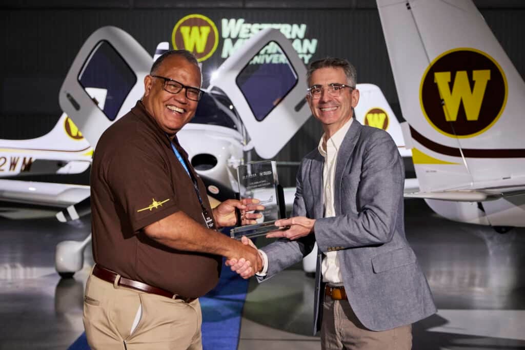 Edward Montgomery, President of the University of Michigan, and Ben Kowalski shake hands in front of a Western Michigan University aircraft inside a hangar. Montgomery, on the left, is wearing a brown shirt with a pilot's badge, and Kowalski, on the right, is in a gray blazer, holding a clear award with text. The Western Michigan University logo is prominently displayed on the tail of the plane in the background.