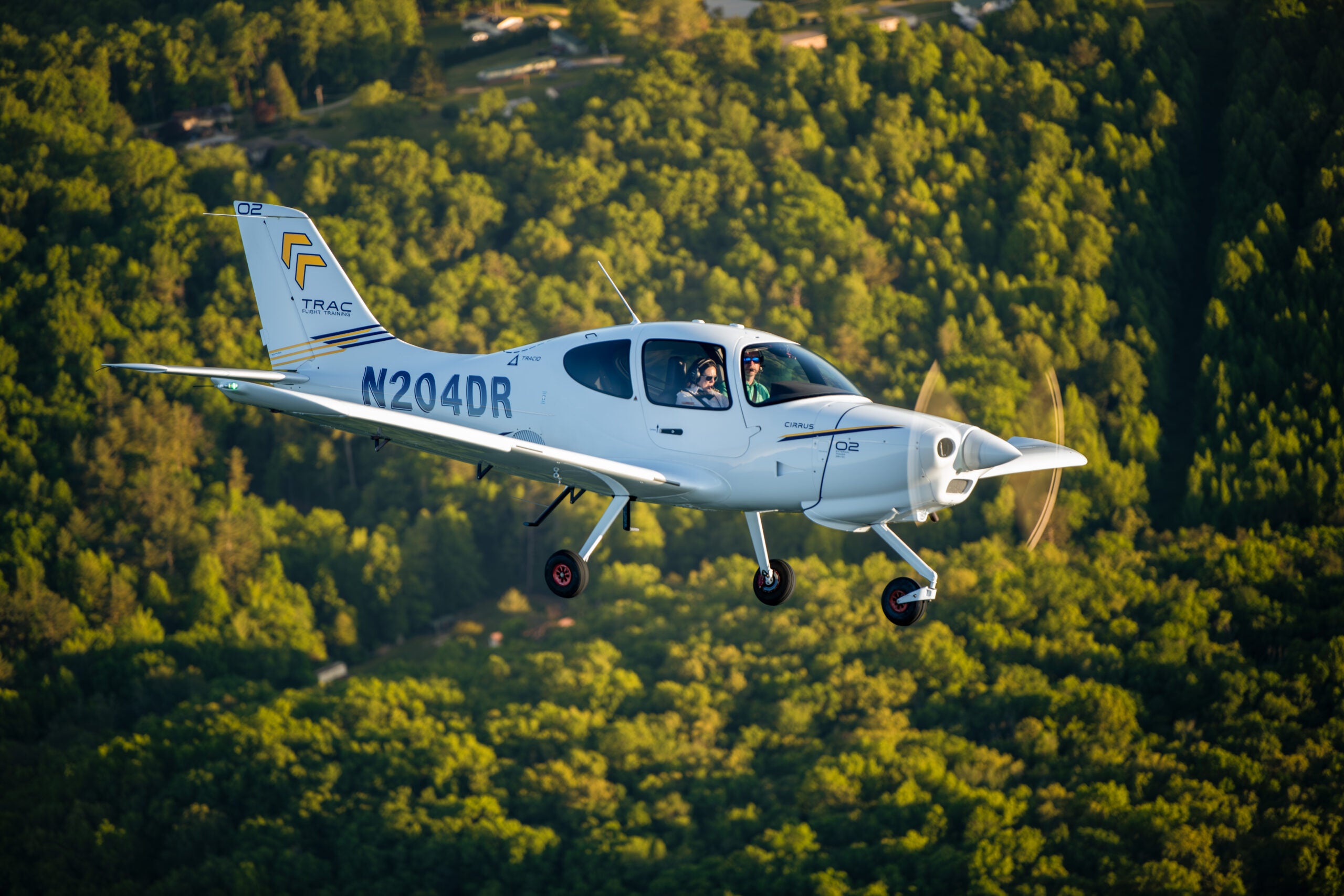 Cirrus TRAC10 flight training aircraft in flight over green forest with an instructor and student aboard
