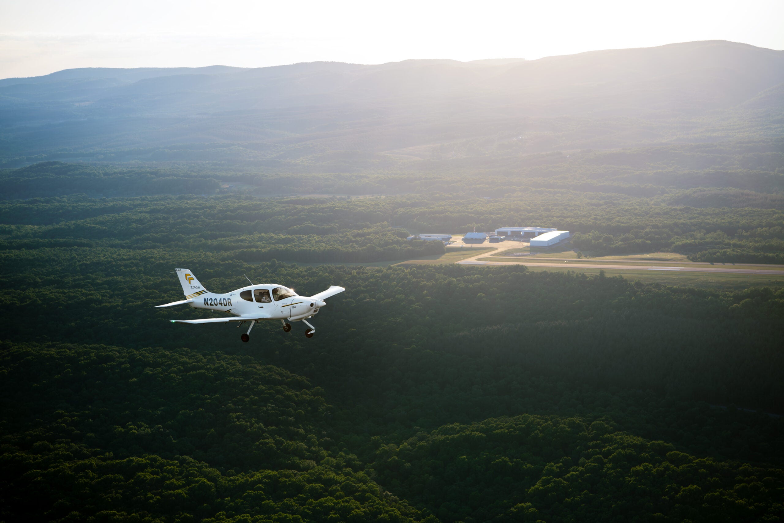 Cirrus TRAC10 training aircraft flying over forested hills with an airport in the distance