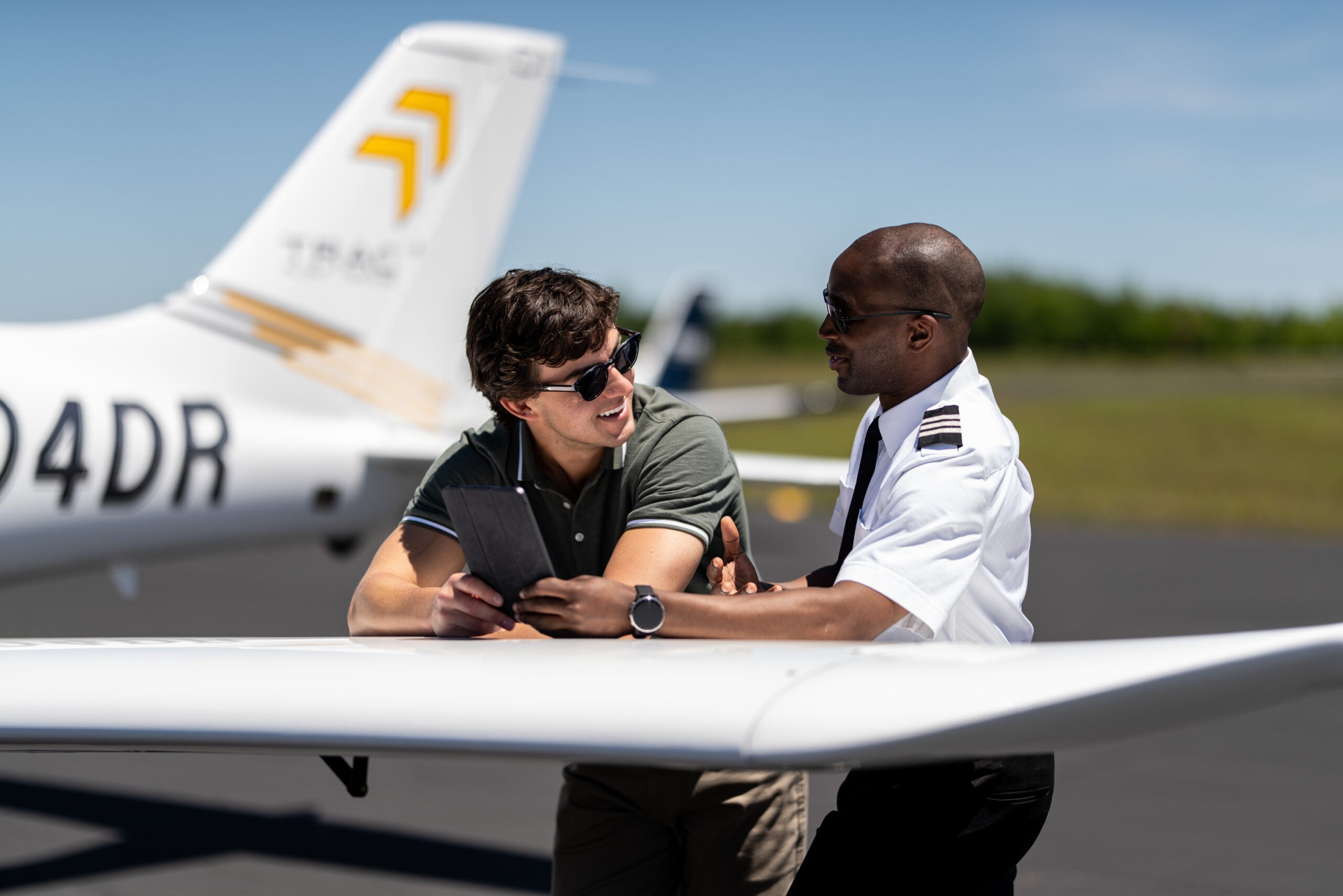 Flight instructor and student pilot reviewing lesson notes on a tablet at the wing of a Cirrus TRAC training aircraft