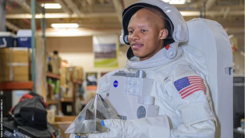 Josh Dobbs wearing a white NASA-style spacesuit with a U.S. flag patch in a training facility