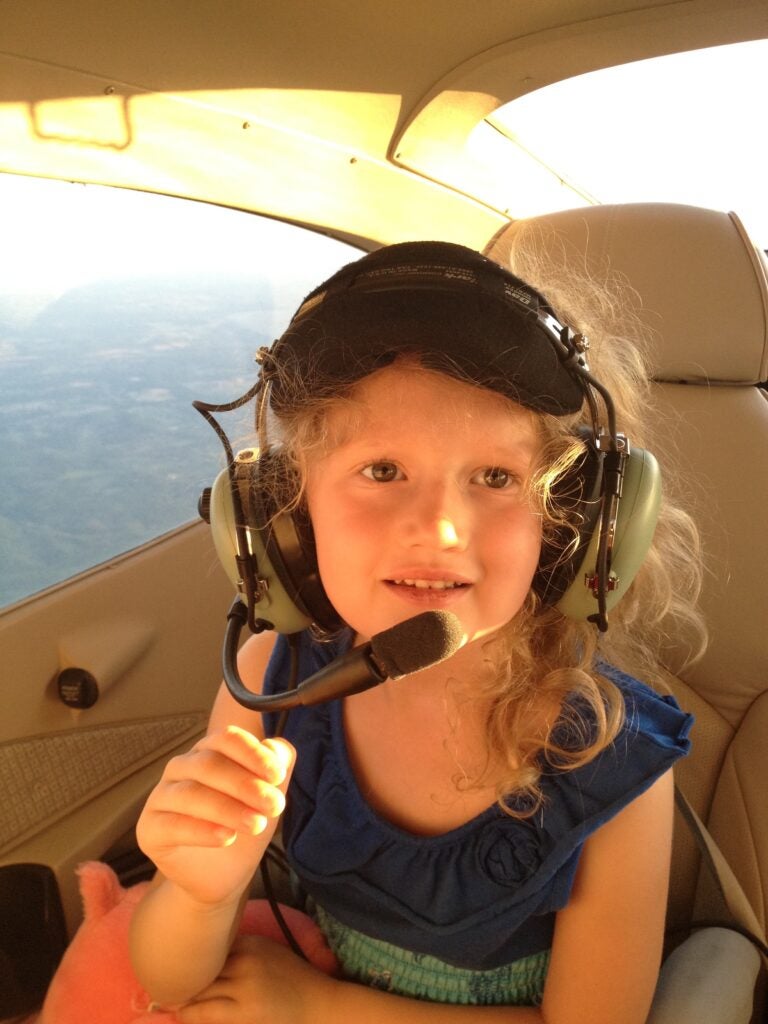 A young Maddie Allen in an aviation headset holding the microphone in the back seat of a small aircraft in flight